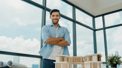 Portrait of architect engineer in casual outfit smile at camera while crossing arms. Businessman looking at camera and standing with arms folded near house model, architectural model. Tracery