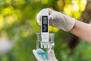 pH meter in hands with gloves, glass of water on blurred background of nature. Measurement of...