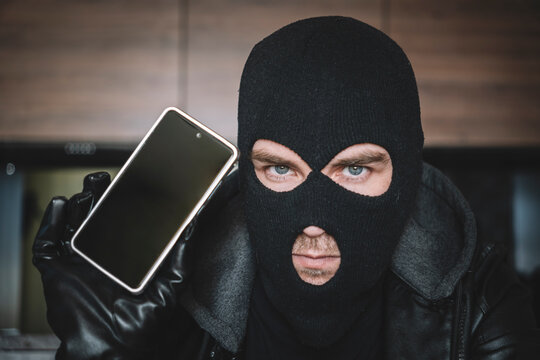 Young adult in black clothes with hidden face. Ill-intended fraudster uses mobile. Fraudster calls. Scam. Mobile racket. Hacker hijacks by phone. Cellphone account fraud.