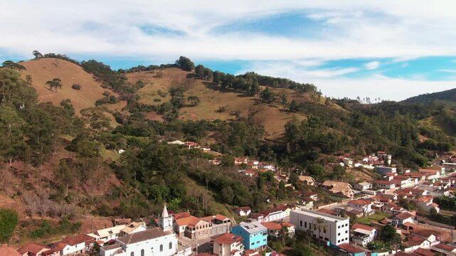 Aerial panorama of Delfim Moreira, Minas Gerais, Brazil, showing a town with red-tiled roofs surrounded by lush green hills and forests.