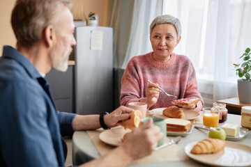 Portrait of Asian senior woman enjoying breakfast at dinner table with husband in cozy kitchen