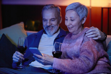 Portrait of smiling senior couple using smartphone sitting on couch in blue light and watching TV