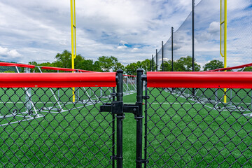 Black chain link fence, gate, with red fence cap and softball / baseball field in background.
