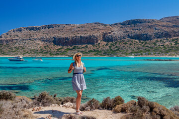 Woman wearing a straw hat is standing on the beach with boats in the background, by the azure water. The scene includes mountains and coastal landforms. Crete. Balos. Greece.