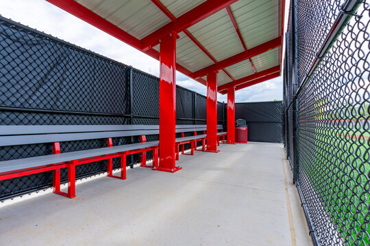 Interior photo of a red softball baseball dugout with roof, concrete floor, and red and gray team bench.  	