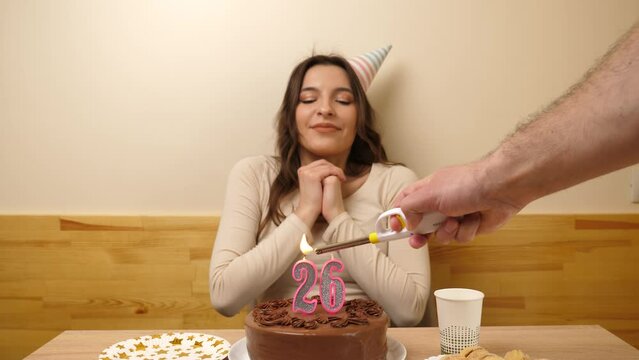 The girl is sitting in front of a table with a festive cake, in which a candle in the form of the number 26 is burning, which she blows out. Birthday celebration concept. Slow motion
