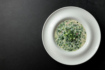 Cold soup with fresh cucumbers, sausage, broth and sour cream in a plate-hat on a black background. A traditional dish is okroshka