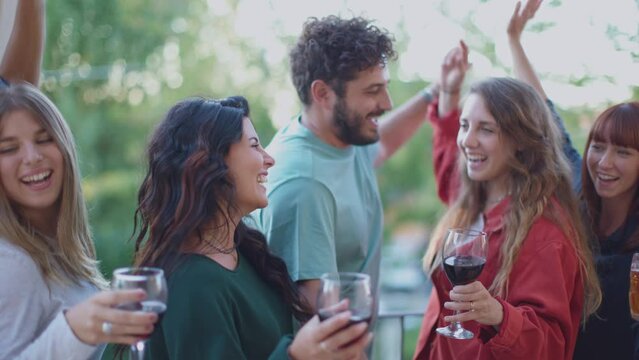 Joyful Generation Z friends dancing and drinking red wine, celebrating a rooftop barbecue party at daylight. Excited young people dancing and having fun outdoors on summer vacation