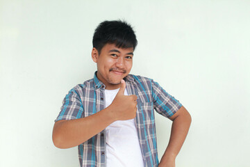 Excited and happy Asian man gives thumbs up hand gesture of approval, isolated by gray background.