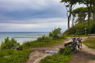 Fototapeta premium Fahrrad-Pause am Ostseeküsten-Radweg mit Blick vom Steilufer auf die Ostsee am Gespensterwald Nienhagen, Fahrräder parken an einer Bank am Kliff vor markanten Buchen unter Wolkenhimmel, Deutschland