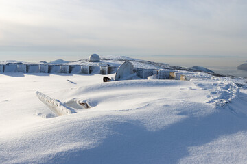 View of the snow-covered ruins and white dome (radio-transparent shelter) of an abandoned radar station on top of a mountain. Marchekanskaya Sopka, Magadan region, Far East of Russia.