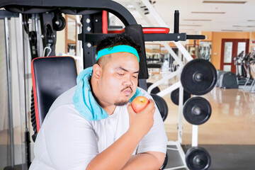 Picture of a young obese man holding a fresh apple while sitting on the exercise machine