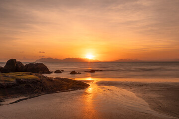 A glowing orange sunrise in a cloudy sky over the sea and beach with reflections in the wet beach...