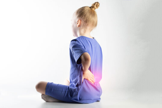 A girl in a purple suit sits and holds her lower back on a white background. Concept of pain and inflammation in the spine in children. Sprains and strains of the back ligaments. Copy space for text