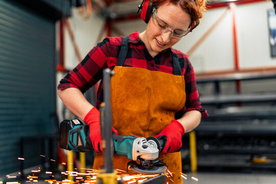 A young girl who is an apprentice in a metal workshop uses a grinder and works on a metal bar, she is wearing protective equipment