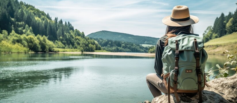 A woman with a backpack and straw hat gazes at a tropical river on a sunny day, with a lush background providing a perfect copy space image.