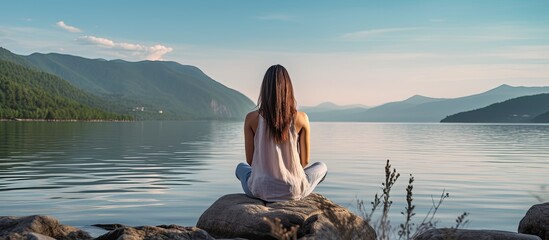 A woman is seen meditating on a hill overlooking the sea in a peaceful setting, with a view from behind, providing ample space for adding text to the image.