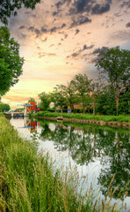 Fototapeta premium Idyllic sunset with beautiful reflections on a tranquil canal, surrounded by greenery and a cloudy sky.