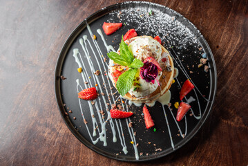 Stack of pancakes with ice cream, mint, and strawberries on a dark plate, drizzled with syrup