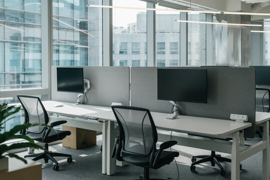 A modern, open-plan office space filled with natural light from large windows that offer a view of the cityscape. The office features a clean, minimalistic design with  chairs and computer monitors.