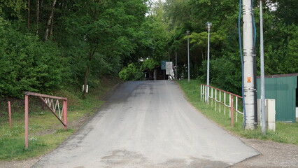 Obraz premium Asphalt Driveway with Open Barrier Gate, Power Pole, and Surrounding Greenery An image featuring an asphalt driveway with an open barrier gate, indicating that access is allowed. A power pole stands n
