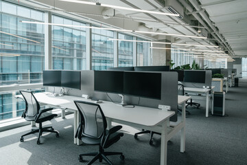 A modern, open-plan office space filled with natural light from large windows that offer a view of the cityscape. The office features a clean, minimalistic design with  chairs and computer monitors.