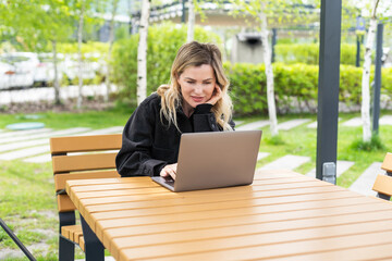 Young woman sitting on online meeting in outdoor cafe, talking to laptop camera, explaining...