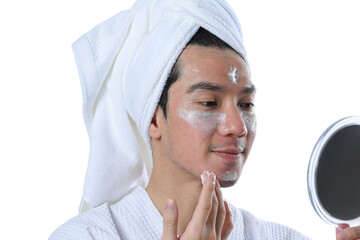 Young man applying moisturizer on his face and looking at mirror over white background