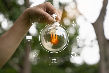 Holding a light bulb against nature on green leaf with energy source icon for renewable and...