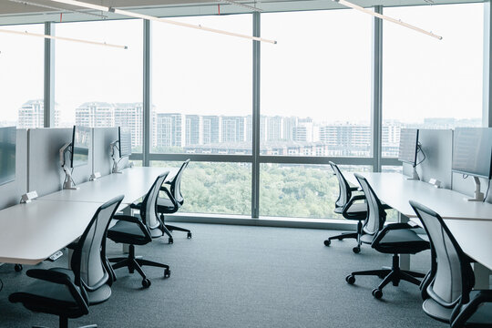 A modern, open-plan office space filled with natural light from large windows that offer a view of the cityscape. The office features a clean, minimalistic design with  chairs and computer monitors.