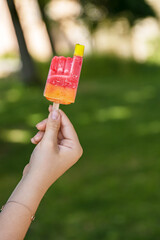 Close-up of a vibrant colorful red yellow orange bitten ice cream with popsicle stick held in a hand of teenager girl. Sunny summer day in park. Blurred background. Copy space. Selective focus