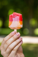 Girl holding and showing bitten yellow red ice cream her blurred face and silhouette are on background. Color frozen fruit popsicle. Hot Sunny day outside. Copy Space. Selective focus