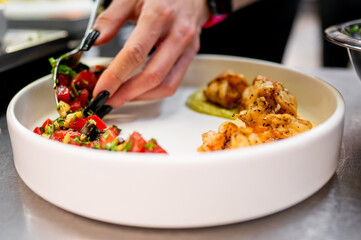 Close-up of a hand garnishing a dish with herbs, featuring grilled shrimp and fresh salad.