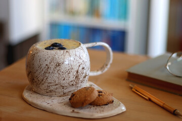 Cup of blueberry and banana smoothie, chocolate chip cookies, book, pen and reading glasses on the table. Colorful bookshelf in the background. Selective focus.