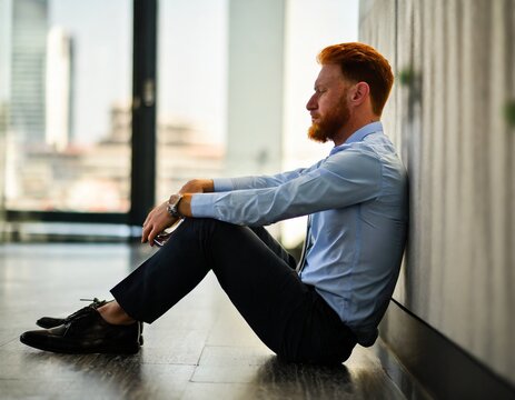 Depressed person sitting on the floor of city office against the wall. Caucasian red haired financial broker. AI generated image.