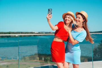 Two young beautiful smiling hipster women in trendy summer same clothes. Carefree models posing in the street. Positive models having fun. Cheerful and happy, at sunny day. In hat. take selfie