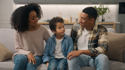 Loving African American ethnic family look at each other male man dad woman female mom little boy child kid son looking camera happy wide smile wave hands hello greeting childcare sit sofa indoor room