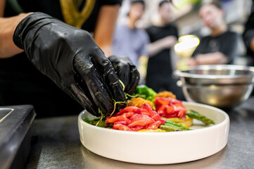 Close-up of hands in black gloves garnishing a fresh salad with red salmon and green vegetables in...