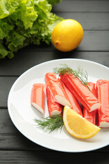 Tasty crab sticks, pieces of lemon and salad on plate on black background.