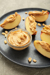 Slices of fresh apple with peanut butter in bowl on blue plate on grey background.