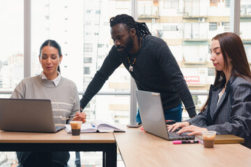 Young African man in office standing looking at laptop of two women sitting working