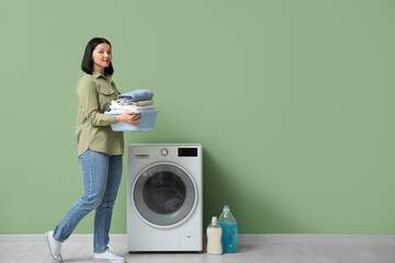 Young woman with laundry walking near washing machine on green background