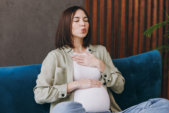 Young pregnant woman wear casual clothes breathing hold hands on belly breast sit on grey sofa couch stay home hotel flat rest relax spend free time in living room indoor. Maternity pregnancy concept.