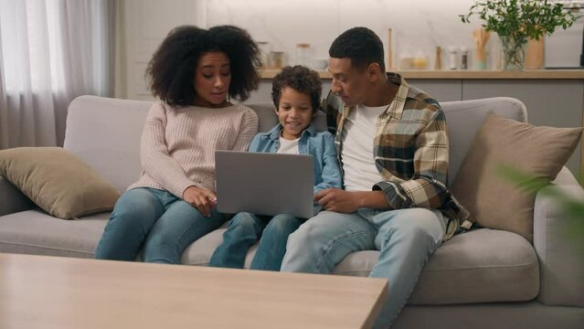 Happy African American family planning vacations travel woman female mom child son kid little boy father male man using laptop notebook discuss talk trip buying tickets online booking indoor room home