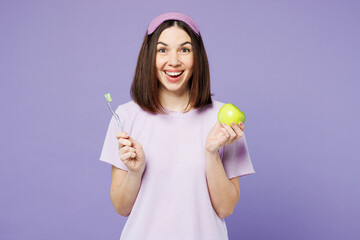 Young surprised shocked happy calm woman wear pyjamas jam sleep eye mask rest relax at home brush teeth hold eat apple isolated on plain pastel purple background studio. Good mood night nap concept.
