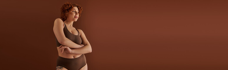 A young, curvy redhead woman in a bikini exudes confidence in front of a brown background.