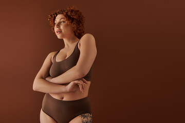 A young, curvy redhead woman poses in a stylish brown bikini against a complementary background,...