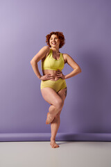 Curvy redhead beauty in yellow bikini striking a pose in front of a vibrant purple wall.