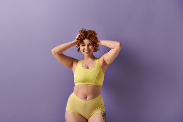 A young, curvy redhead woman confidently poses in a yellow bikini against a striking purple...