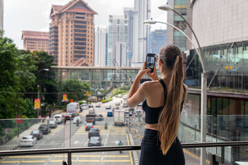 A woman in a black top photographs the Kuala Lumpur skyline from a glass bridge, capturing...